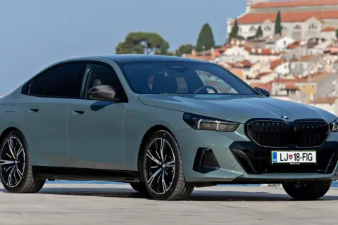 Szary sedan BMW 5 2025 z czarnym grillem i felgami, zaparkowany na tle malowniczego miasta nad morzem.