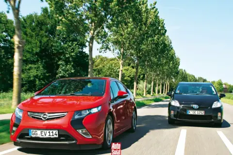 Czerwony Opel Ampera i czarny samochód jadą drogą otoczoną drzewami.