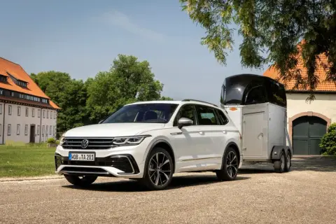 Biały Volkswagen Tiguan, jeden z wielu wszechstronnych volkswagen modele, holuje przyczepę z koniem przed zabytkowym budynkiem.