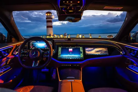 Wnętrze luksusowego mercedesa e220d oświetlone nastrojowym, wielokolorowym ambientowym światłem. Widok na morze i latarnię morską przez przednią szybę.