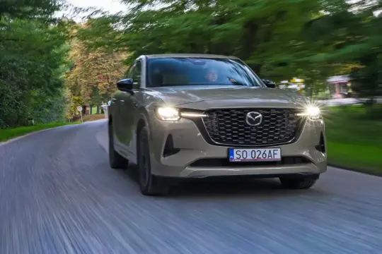 Elegancka Mazda CX-60 3.3 diesel w kolorze piaskowym pokonuje zakręt na leśnej drodze, światła LED oświetlają asfalt.