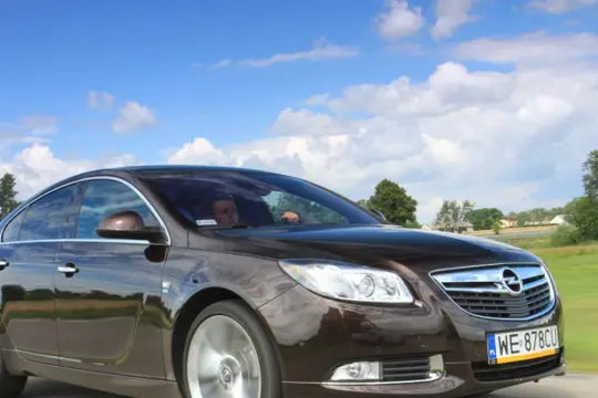 Brązowy Opel Insignia 2.0 CDTI w ruchu, z kierowcą za kierownicą, na tle błękitnego nieba z chmurami i zielonych pól.