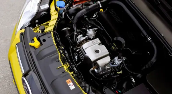 Silnik 1.0 Ecoboost w żółtym Fordzie. Widoczny jest czarny kolektor dolotowy z napisem Ecoboost, elementy metalowe i przewody.