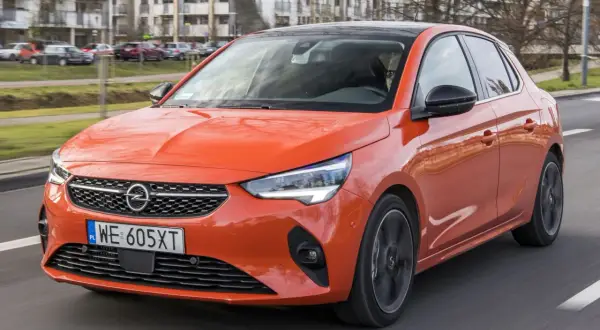 Pomarańczowy Opel Corsa 1.2 z czarnym dachem jedzie po drodze, w tle budynki mieszkalne.