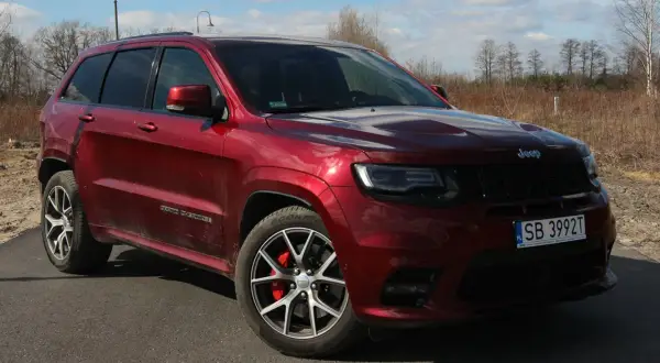 Czerwony Jeep Grand Cherokee SRT z czarnymi felgami i czerwonymi zaciskami hamulcowymi, zaparkowany na drodze wśród suchych traw i drzew.