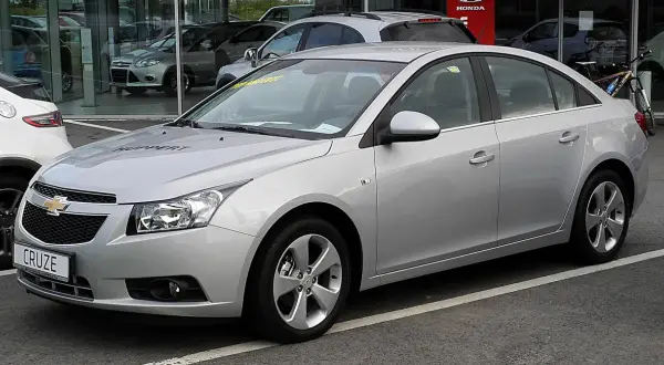 Srebrny Chevrolet Cruze 1.8 benzyna z napisem "HUPPERT" na masce, zaparkowany przed salonem samochodowym.
