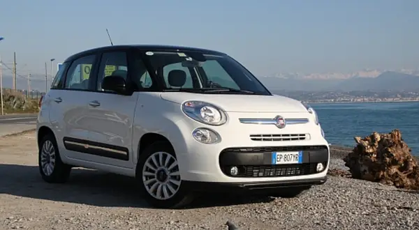 Biały fiat 500 l z czarnym dachem stoi na kamienistej plaży nad morzem.