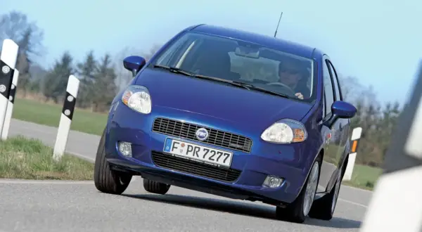 Niebieski Fiat Punto Grande pokonuje zakręt na drodze, z kierowcą w okularach za kierownicą.
