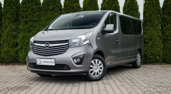 Szary Opel Vivaro, jeden z popularnych modeli Opla, zaparkowany przed żywopłotem.