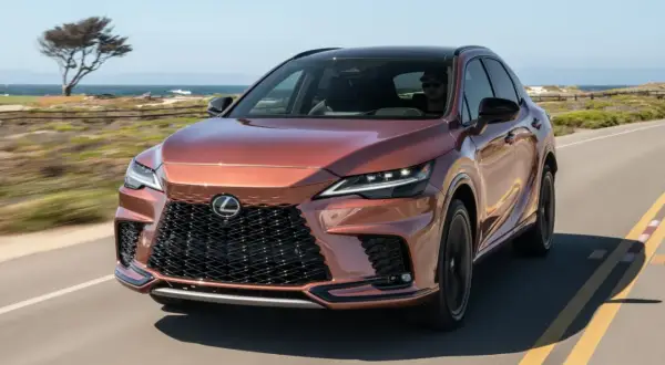 Miedziany Lexus RX 500h jedzie wzdłuż wybrzeża. Słońce odbija się od jego lśniącej karoserii.