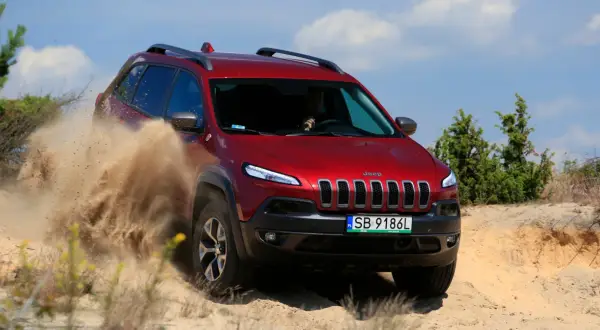 Czerwony Jeep Cherokee Trailhawk z impetem przemierza piaszczysty teren, wzbijając tumany piasku.