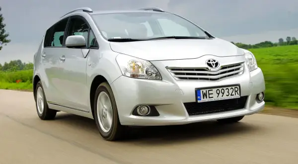 Srebrny Toyota Verso S jedzie po drodze, otoczony zielonymi polami.
