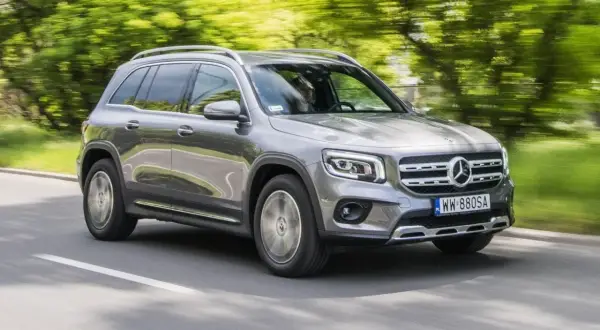 Szary Mercedes GLB 200 jedzie po drodze, otoczony zielenią drzew.
