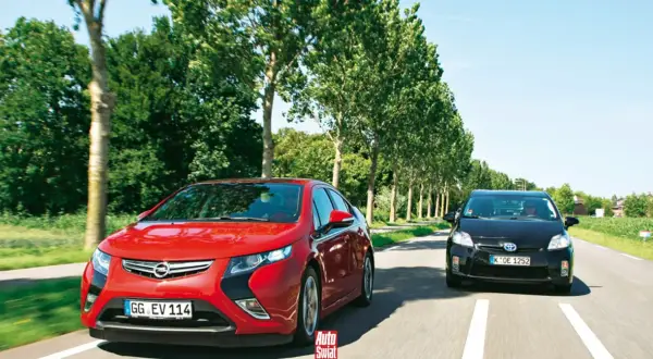 Czerwony Opel Ampera i czarny samochód jadą drogą otoczoną drzewami.