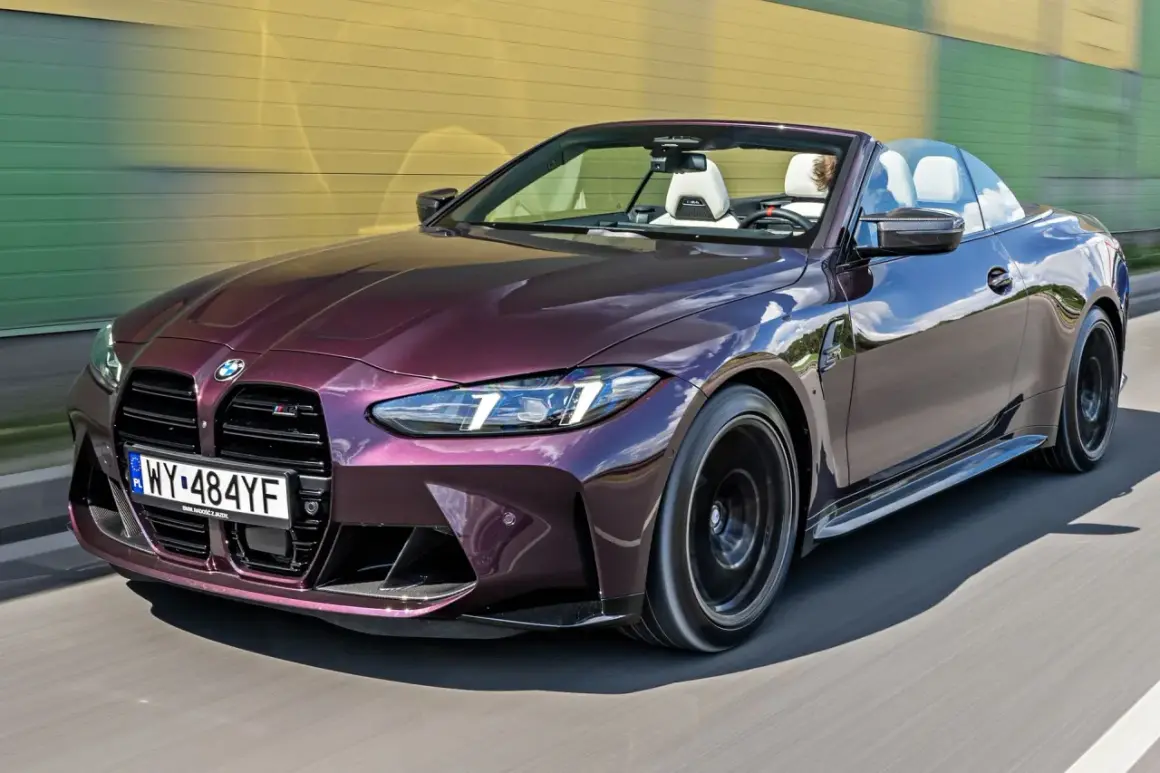 Fioletowy kabriolet BMW M4 F82 z białym wnętrzem pędzi po drodze, odbijając niebo i żółto-zieloną ścianę.