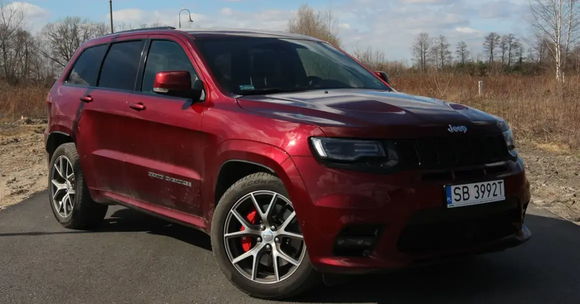 Czerwony Jeep Grand Cherokee SRT z czarnymi felgami i czerwonymi zaciskami hamulcowymi, zaparkowany na drodze wśród suchych traw i drzew.