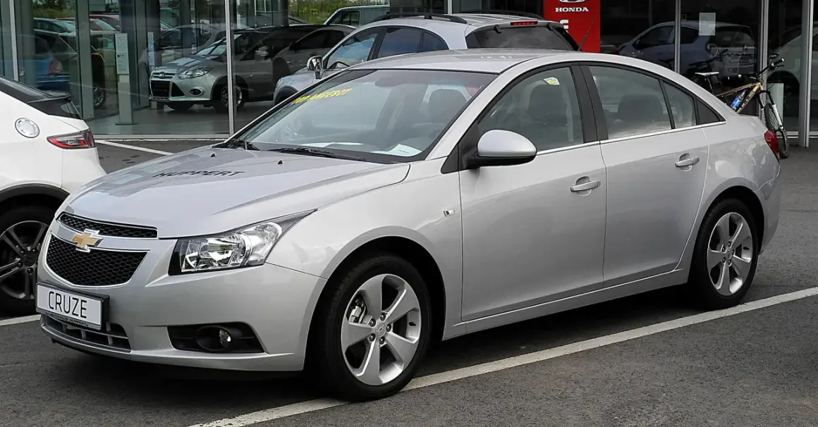 Srebrny Chevrolet Cruze 1.8 benzyna z napisem "HUPPERT" na masce, zaparkowany przed salonem samochodowym.
