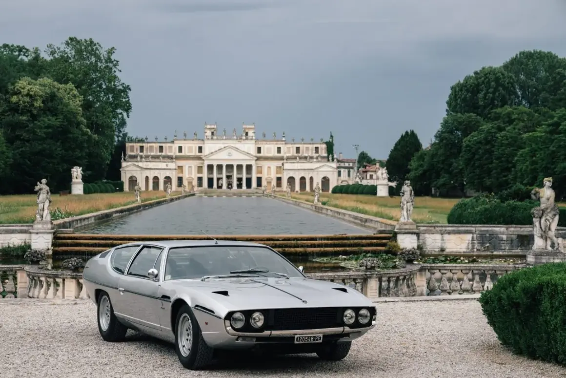 Srebrny Lamborghini Espada przed okazałą willą z basenem i rzeźbami. Klasyk motoryzacji w malowniczej scenerii.