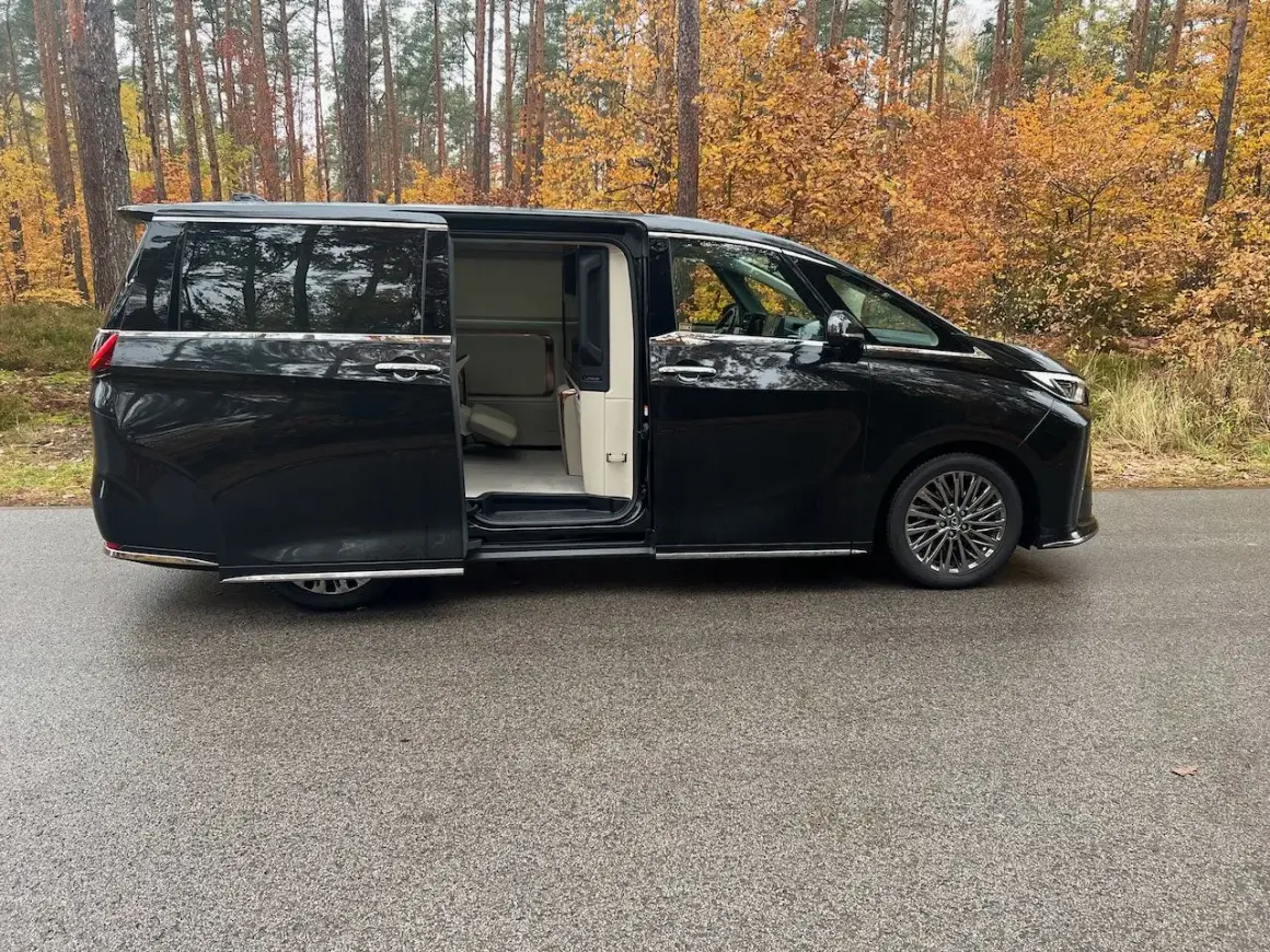 Lexus LM 350h w czarnym kolorze, z otwartymi drzwiami, prezentuje luksusowe wnętrze. Otoczony jesiennym lasem.