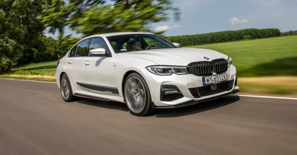 Białe BMW 320d xDrive pędzi po drodze, otoczone zielenią pól i drzew.