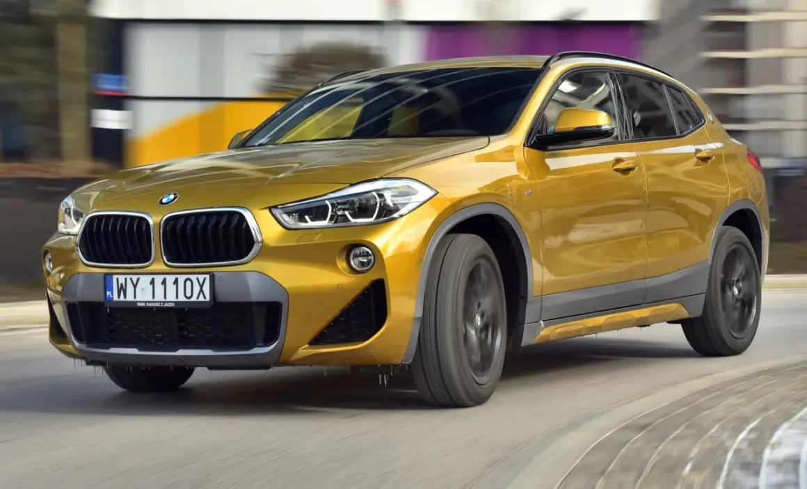 Złoty BMW F39 w ruchu, z widocznym przodem i bokiem, na tle rozmytego miejskiego krajobrazu.