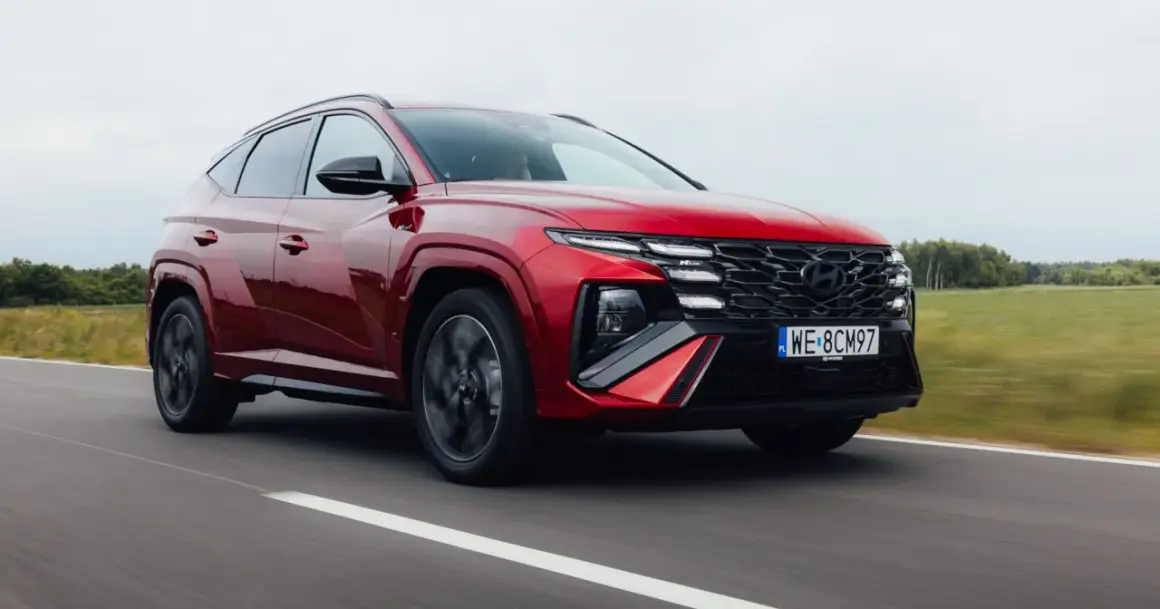 Czerwony Hyundai Tucson 1.6 T-GDI N Line pędzi po drodze, otoczony zielonymi polami i drzewami.