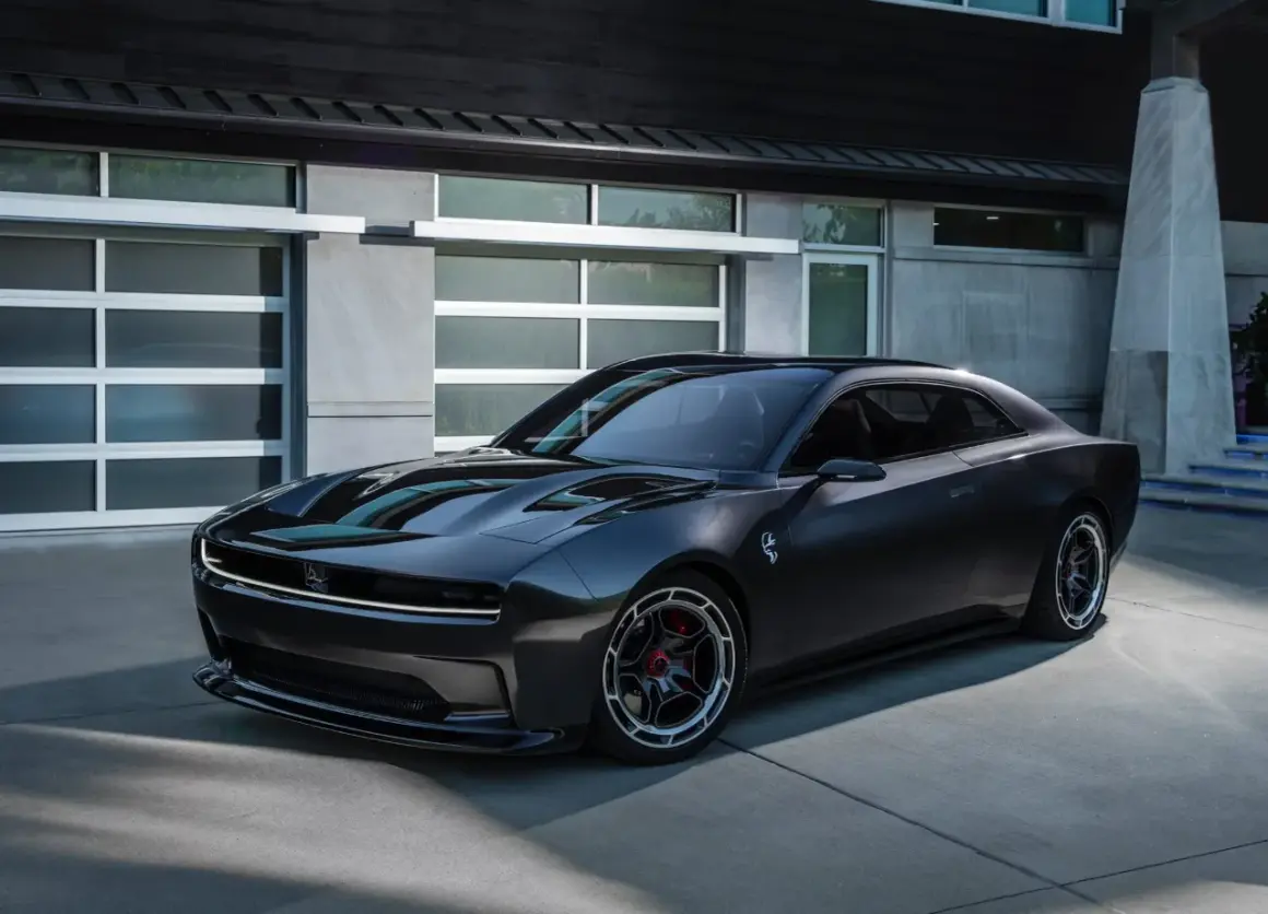 Nowoczesny, ciemnoszary Dodge Daytona z charakterystycznym pasem LED z przodu, zaparkowany przed nowoczesnym budynkiem z garażem.