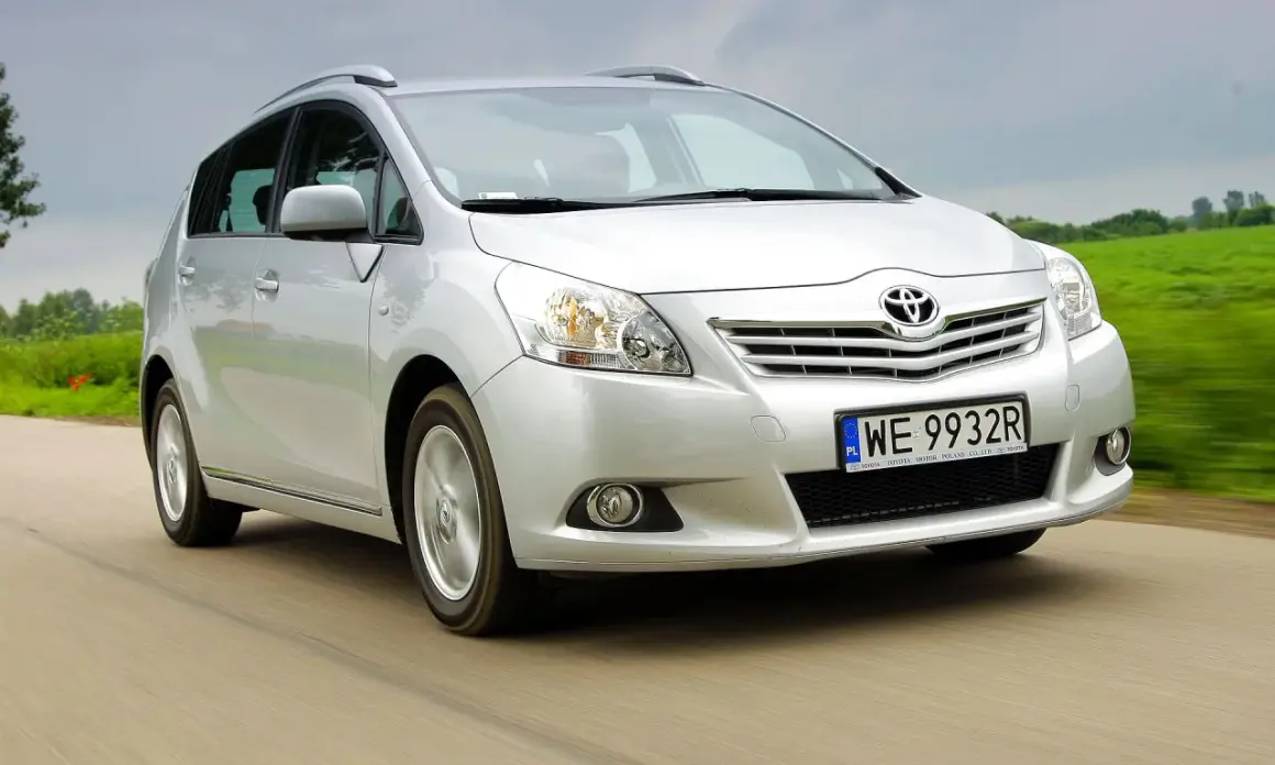 Srebrny Toyota Verso S jedzie po drodze, otoczony zielonymi polami.