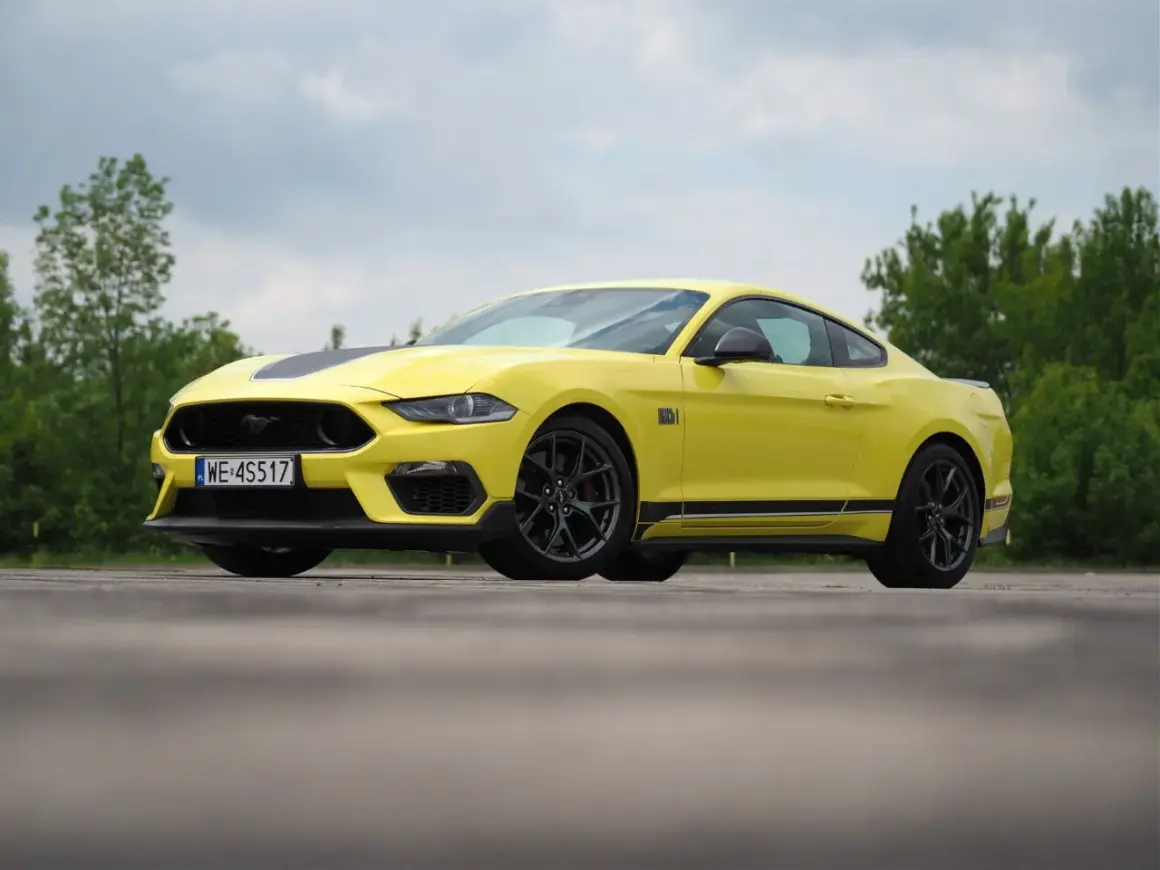 Żółty Ford Mustang Mach 1 z czarnymi pasami na masce i bokach, zaparkowany na tle zieleni.