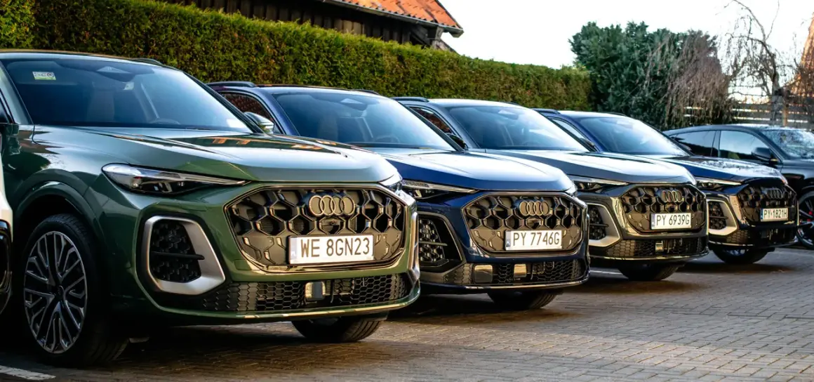 Rząd nowych Audi Q5 w różnych kolorach, od zielonego po ciemne odcienie, zaparkowanych na zewnątrz.