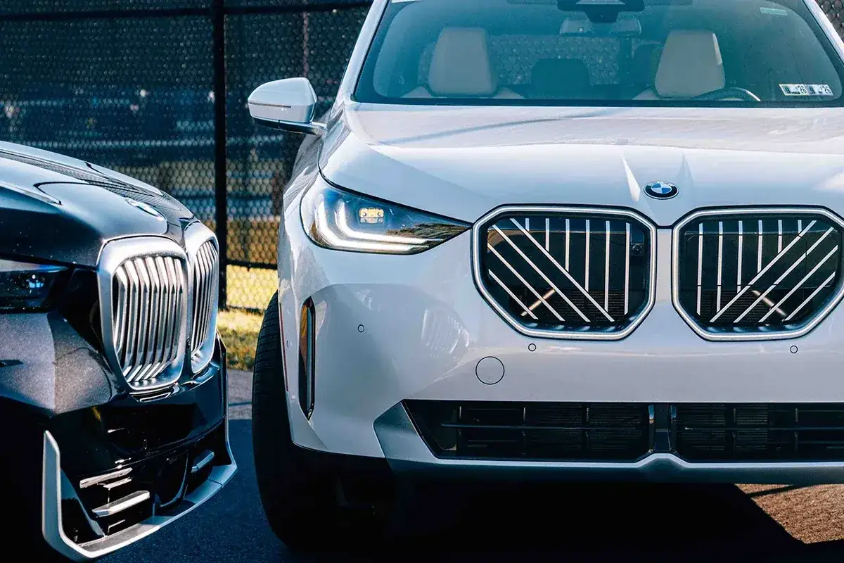 Przód białego BMW X5 2026 z charakterystyczną atrapą chłodnicy i nowoczesnymi światłami. Obok czarny, luksusowy sedan.