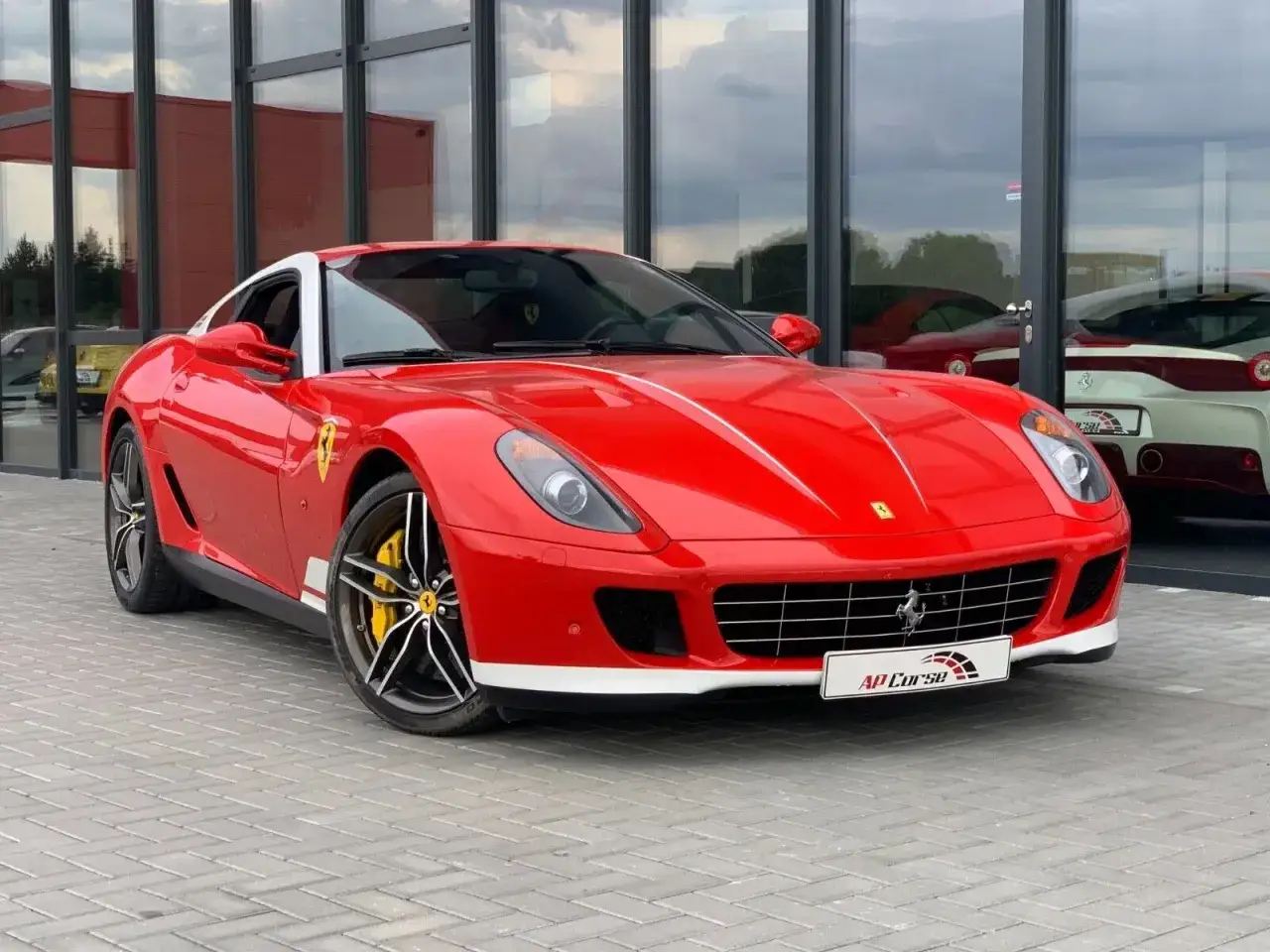 Czerwone Ferrari 599 z białym dachem i żółtymi zaciskami hamulcowymi, zaparkowane przed nowoczesnym budynkiem z lustrzanymi oknami.