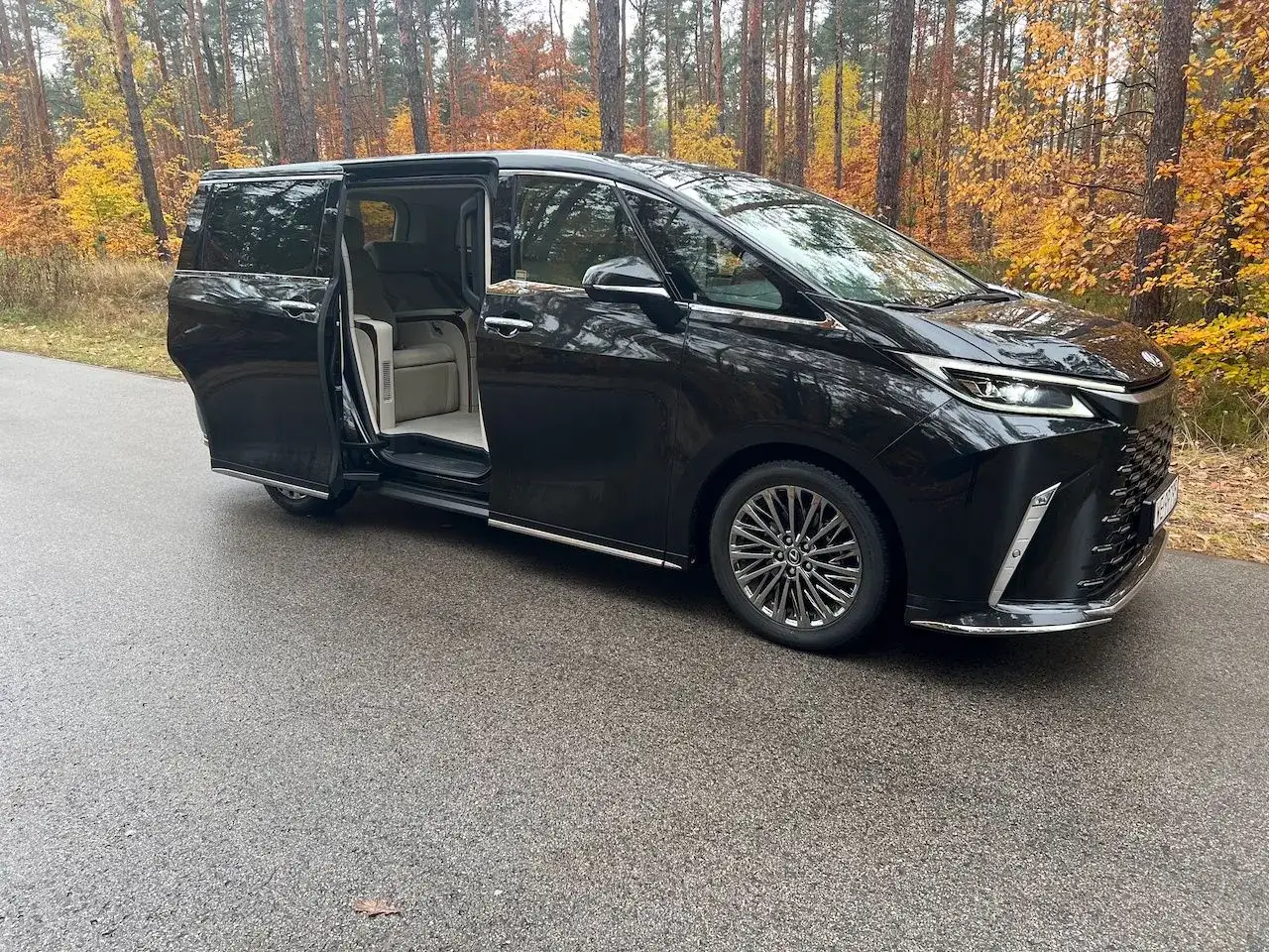 Lexus LM 350h z otwartymi drzwiami, zaparkowany na mokrej drodze w jesiennym lesie.
