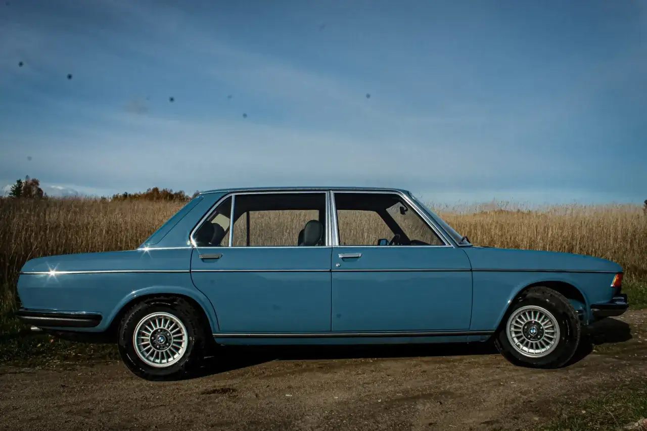 Błękitne BMW E3 na tle jesiennego krajobrazu. Klasyk z lat 70. na drodze wśród traw.