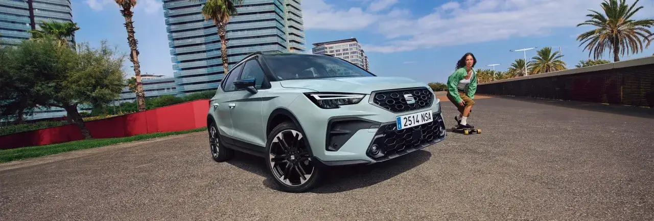 Jasnoszary SEAT Arona, jeden z modeli SEAT, parkuje obok młodej kobiety na deskorolce, na tle nowoczesnych budynków i palm.