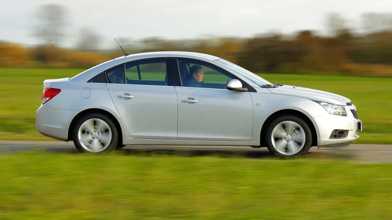 Srebrny Chevrolet Cruze 1.8 benzyna w ruchu, z kierowcą za kierownicą, przemierza wiejską drogę wśród zielonych pól.