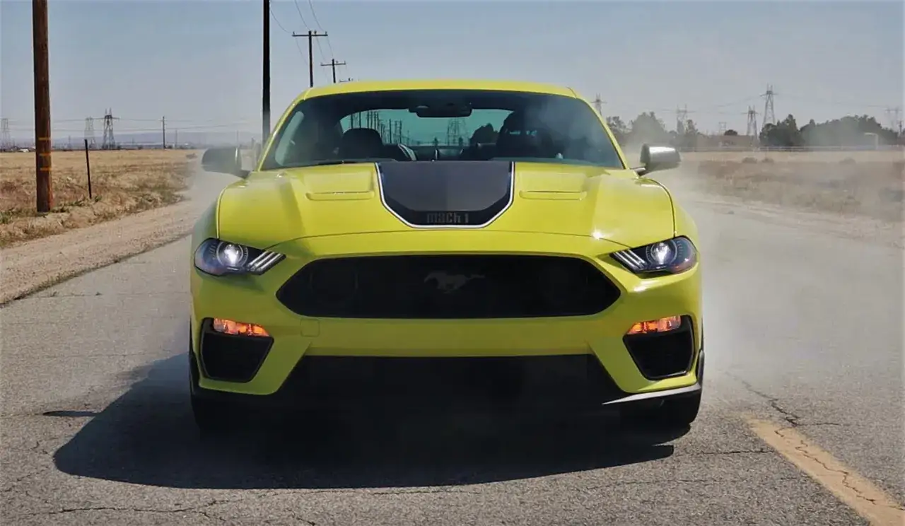 Żółty Ford Mustang Mach 1 pędzi po pustej drodze, wzbijając tumany kurzu.