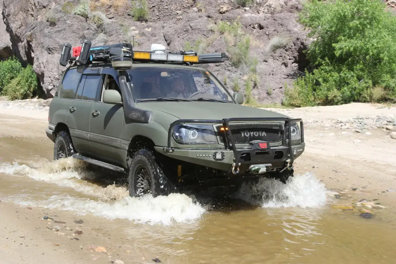 Zielony Land Cruiser 100 pokonuje wodę w skalistym terenie.