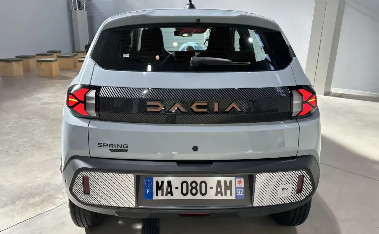 Tył jasnoszarej dacia elektryczna Spring. Litery DACIA w kolorze miedzianym na czarnym pasie.