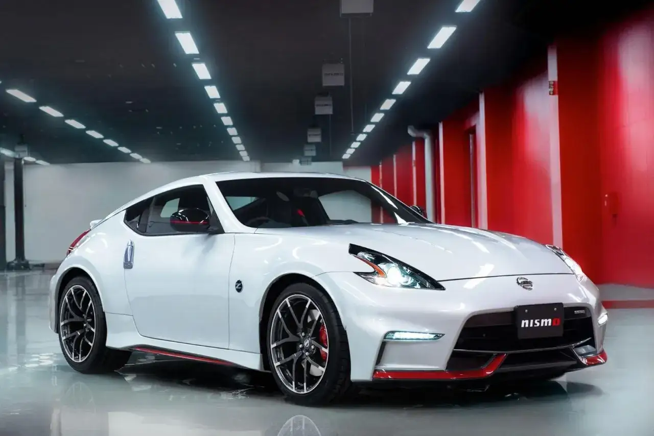 Biały Nissan 370Z Nismo z czerwonymi akcentami, lśniący w garażu z czerwonymi ścianami i sufitowym oświetleniem.