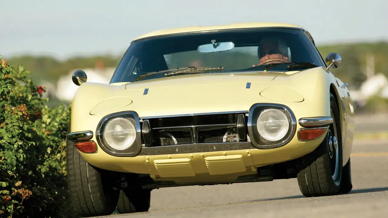 Żółta Toyota 2000GT z lat 60. z kierowcą za kierownicą, jadąca po drodze obok krzewów.