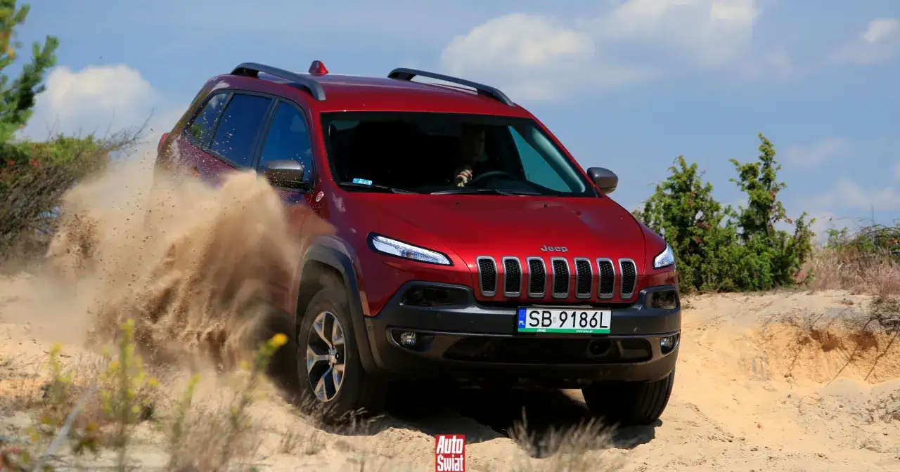 Czerwony Jeep Cherokee Trailhawk szaleje po piaszczystym terenie, wzbijając tumany kurzu.