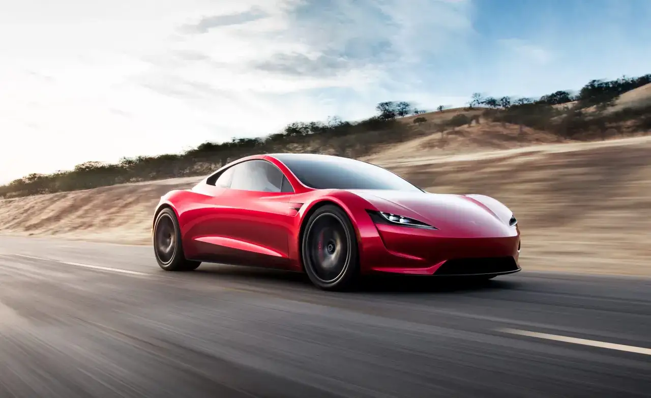 Czerwony Tesla Roadster pędzi po pustej drodze, otoczony suchymi wzgórzami i błękitnym niebem.