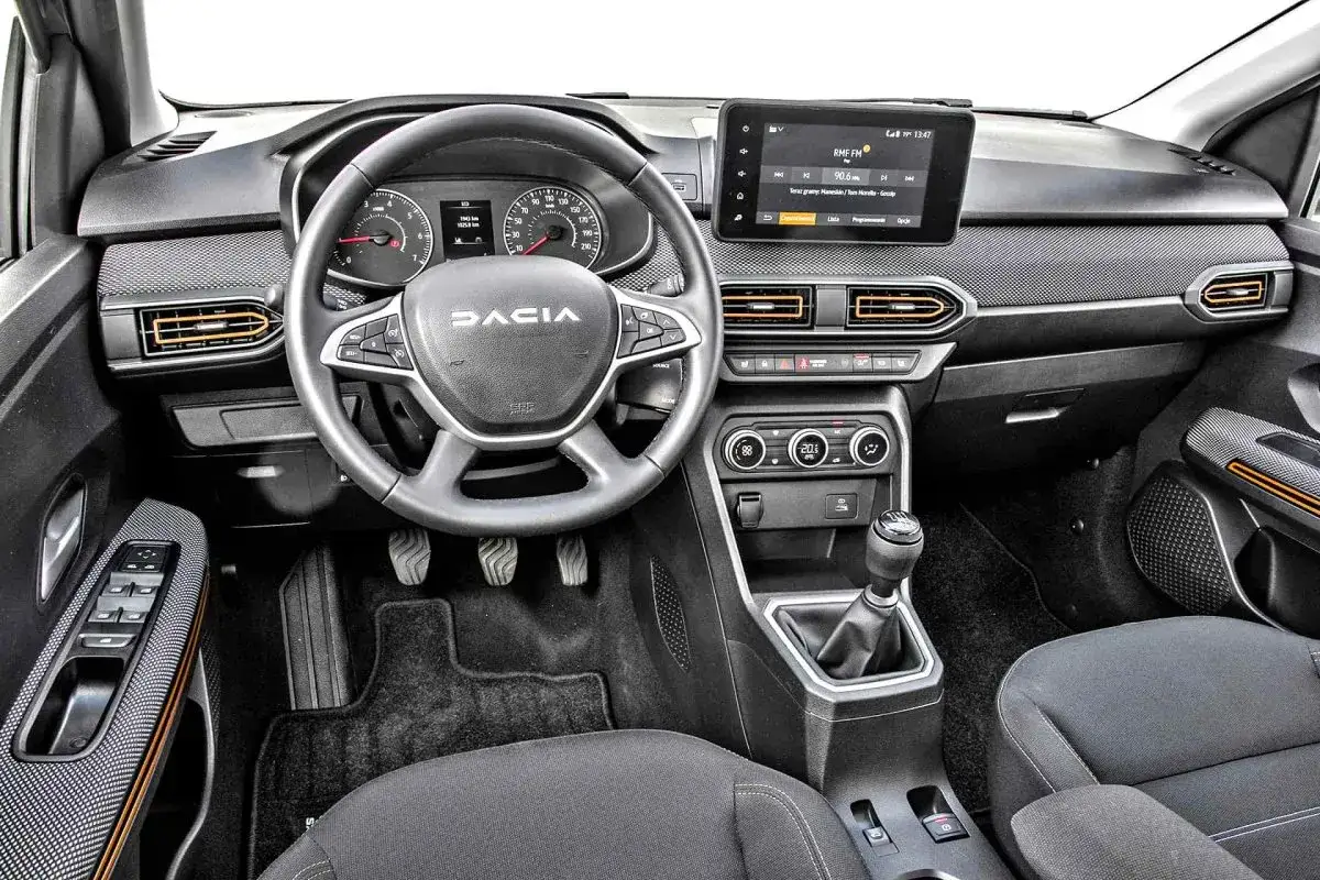 Wnętrze nowej Dacii Sandero: kierownica z logo, ekran multimedialny, panel klimatyzacji i dźwignia zmiany biegów.