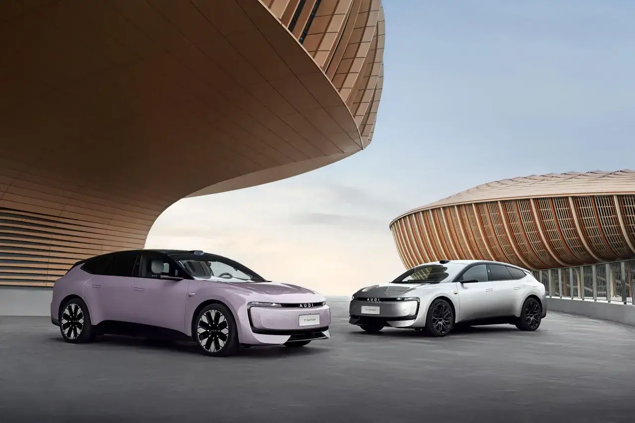 Dwa najnowsze Audi, jedno w kolorze lawendowym, drugie srebrne, stoją przed nowoczesnymi budynkami o falujących kształtach.