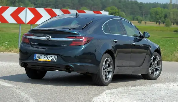 Ciemnozielony Opel Insignia 1.6 Turbo jedzie po drodze, za nim widać znaki ostrzegawcze i zielone pola.