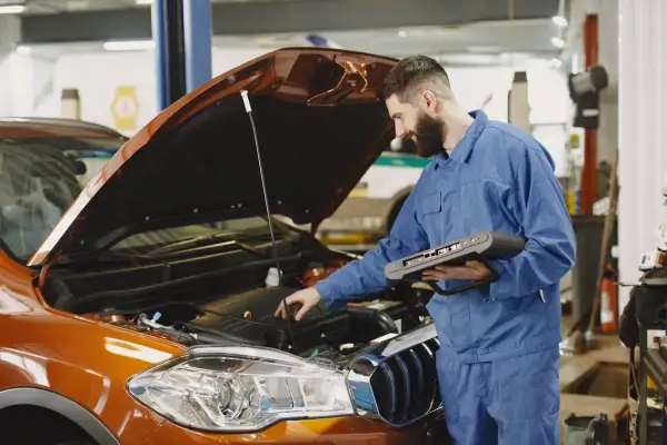Mechanik w niebieskim kombinezonie sprawdza silnik pomarańczowego samochodu za pomocą urządzenia diagnostycznego.
