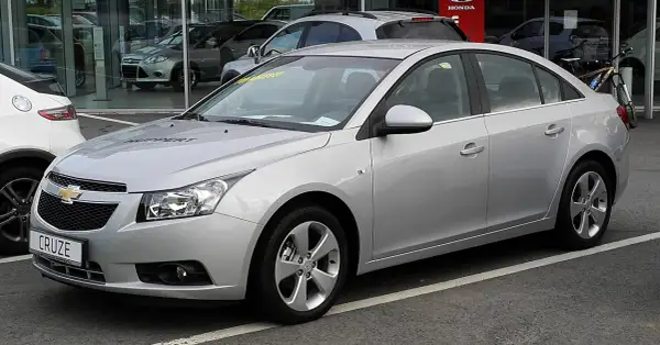 Srebrny Chevrolet Cruze 1.8 benzyna z napisem "HUPPERT" na masce, zaparkowany przed salonem samochodowym.